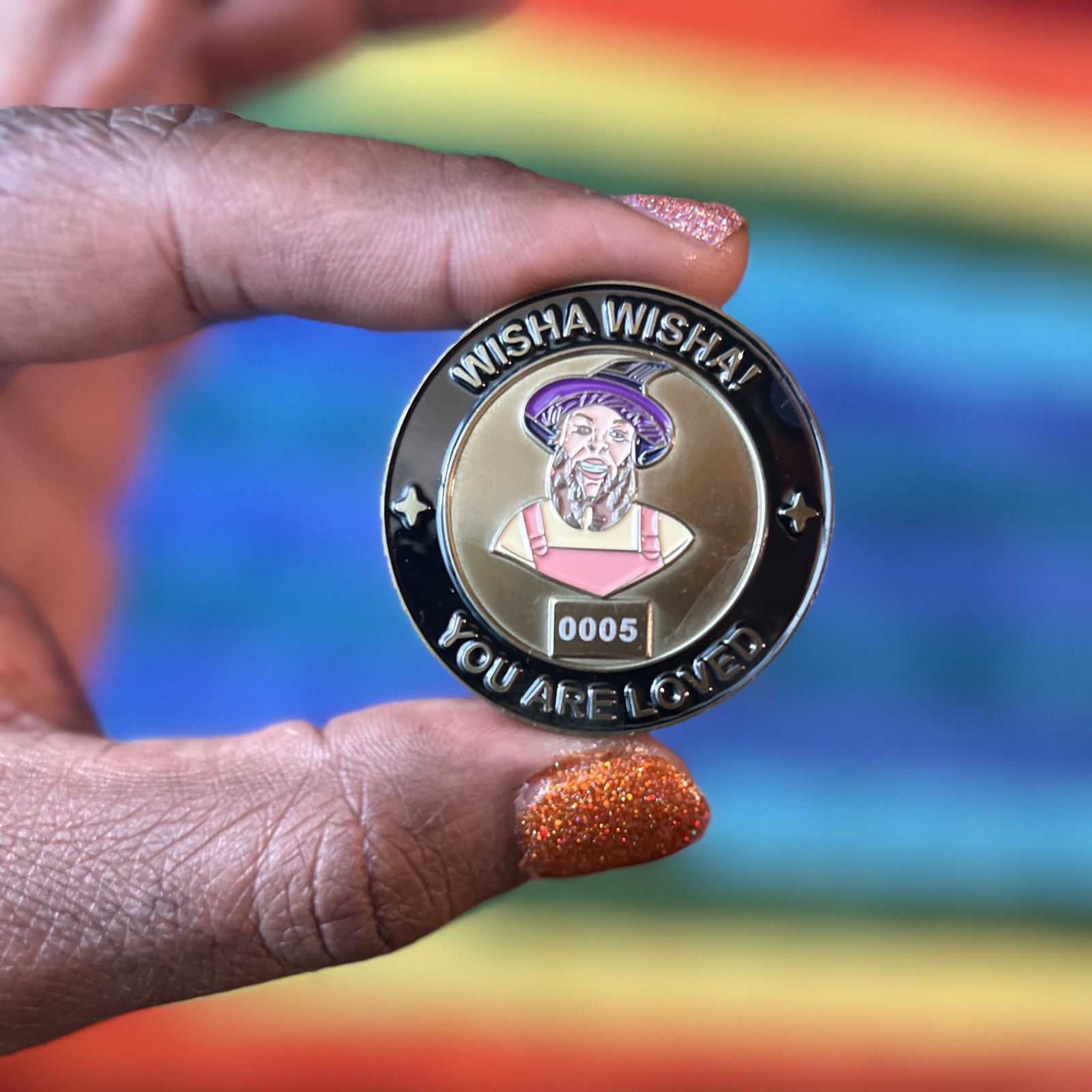 A hand with glittery orange nail polish holds Wizard Coin number 0005, side A, in front of a rainbow pride flag. The coin shows Gilly in a purple wizard hat with the words WISHA WISHA! on top and YOU ARE LOVED below.