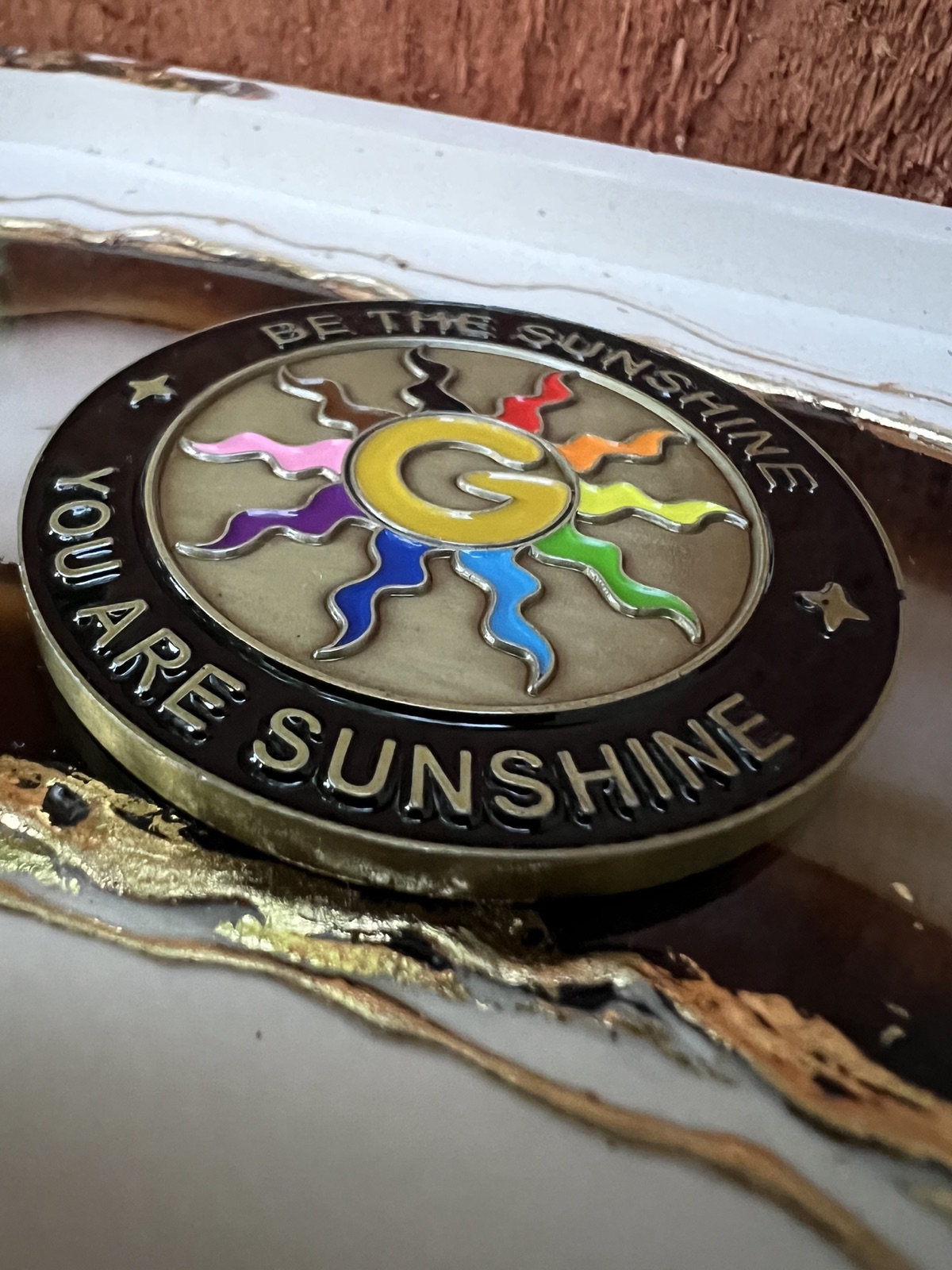 Closeup of the wizard coin's B side resting on a gold-edged agate tray. A sunny yellow letter G with ten colorful squiggly sun rays, ringed by the words BE THE SUNSHINE and YOU ARE SUNSHINE.